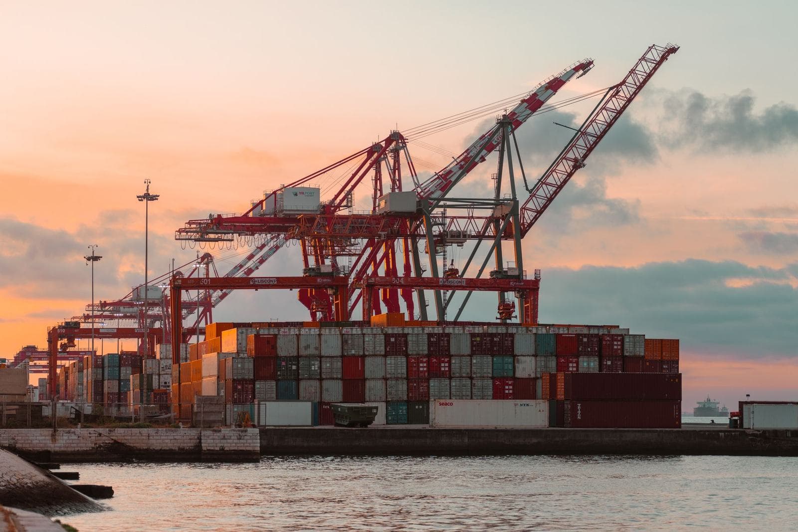 Ship-to-shore cranes over stacked containers at a waterfront terminal, silhouetted against a sunset sky