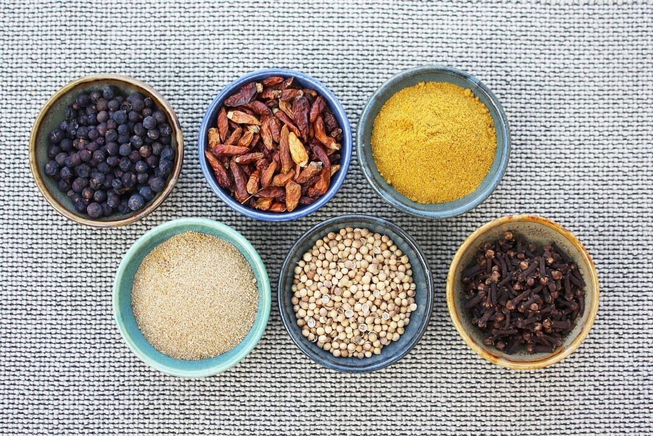Whole and ground spices — peppercorns, dried chillies, turmeric, coriander seed and cloves — in six small bowls on linen