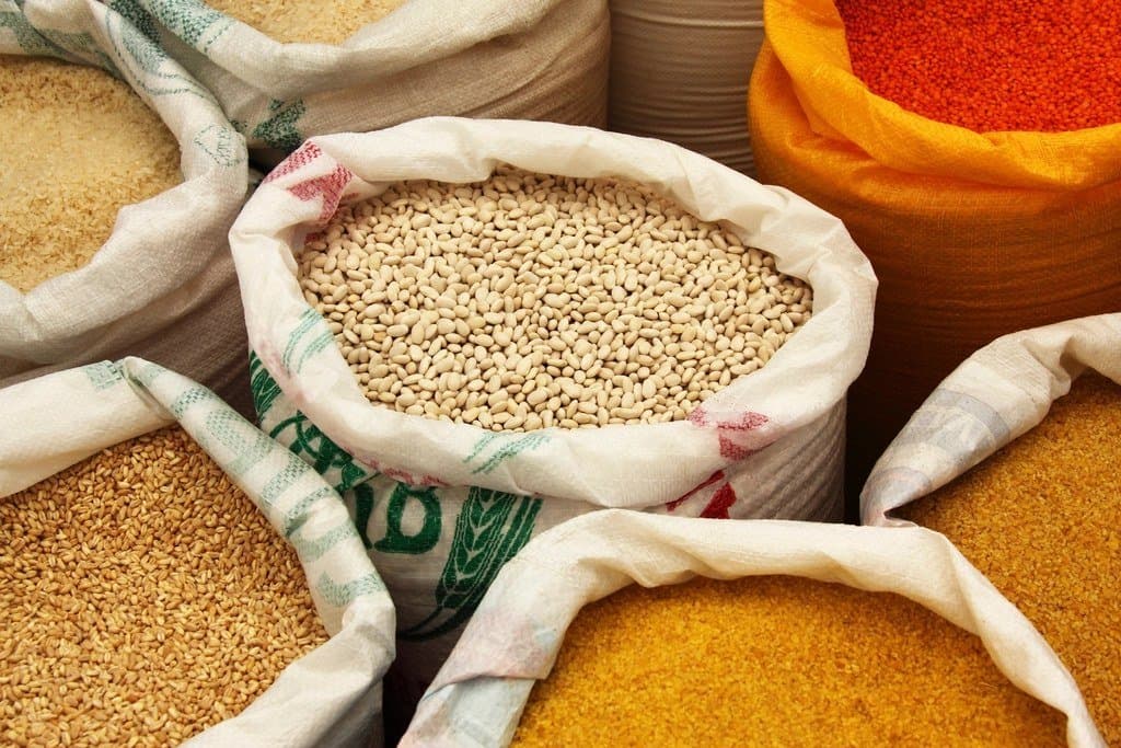 Open woven bulk sacks of cereals and pulses — beans, wheat, rice and lentils — standing side by side on a packing bench