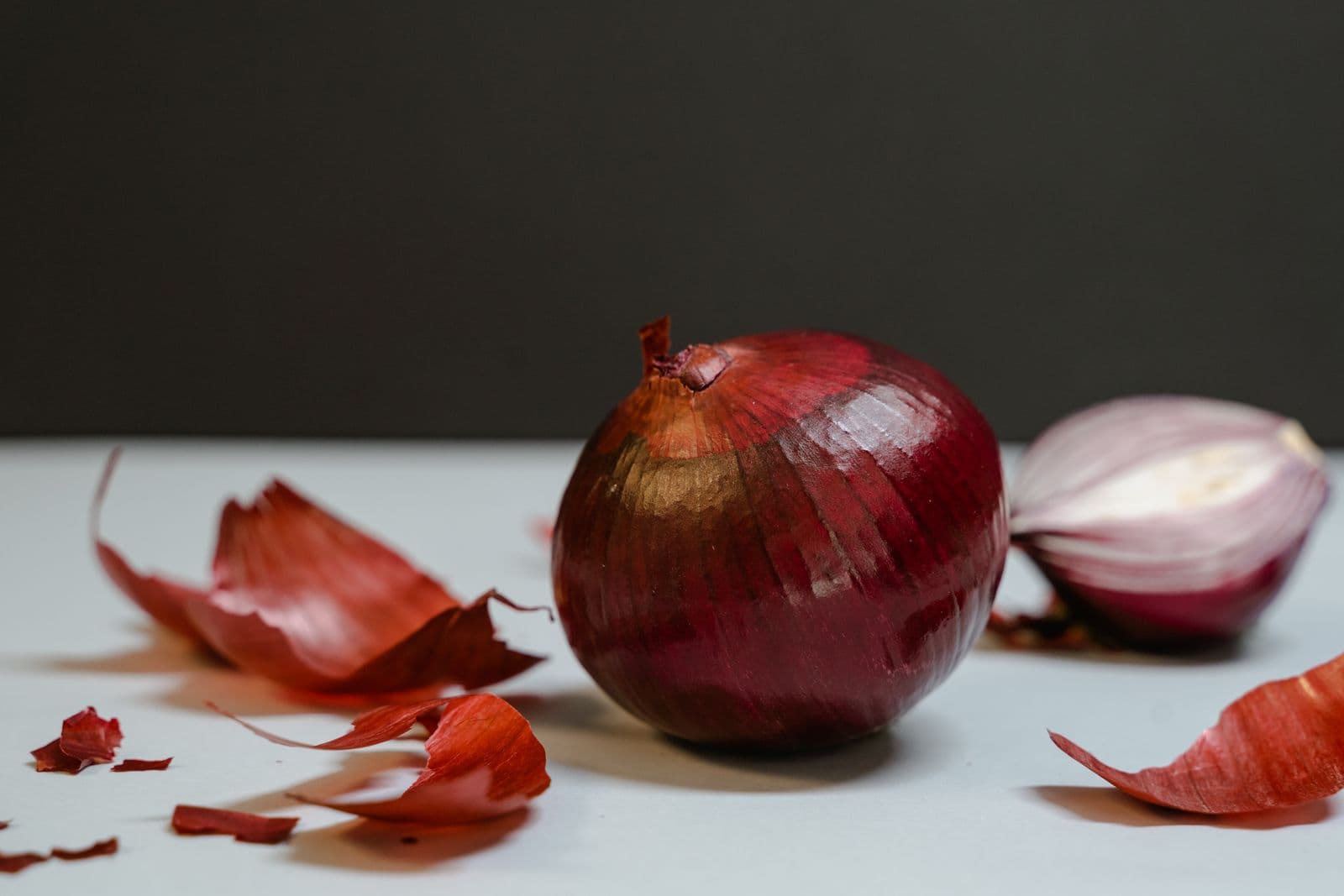 A whole red onion beside a halved one, with loose papery skins on a pale surface
